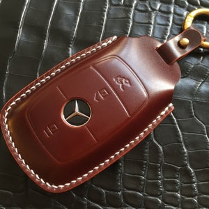 May include: A brown leather key fob with white stitching detail and a silver Mercedes-Benz logo. The fob has a gold metal key ring attached.