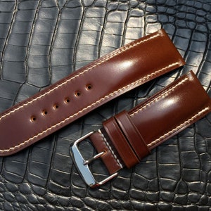 May include: Two brown leather watch straps with white stitching. The straps are laid out on a black alligator-embossed surface.