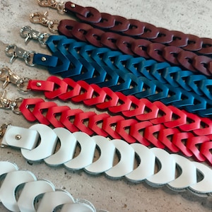 May include: A collection of five braided bag straps in dark brown, deep blue, bright red, and two shades of white. Each strap features a woven leather design and is finished with a polished silver-tone metal swivel lobster clasp at the end.