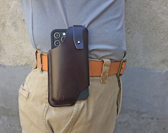 May include: A dark brown leather phone holster attached to a tan leather belt. The holster features a vertical strap with a brass-coloured metal snap closure and a small embossed cat icon on the lower left corner, holding a black smartphone.