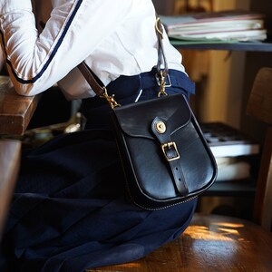 May include: A black leather crossbody bag with a brass buckle and a flap closure. The bag has a strap that goes over the shoulder and a smaller pocket on the front.