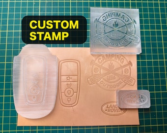 Könnte beinhalten: Vier transparente, kundenspezifische Stempel auf einer grünen Schneidematte. Die Stempel sind für einen Autoschlüssel, ein Camping-Logo und das Land Rover-Logo. Die Stempel befinden sich auf einem Stück braunem Leder.