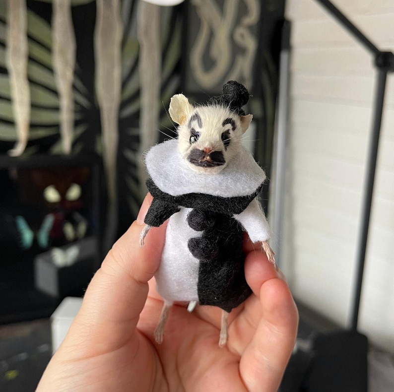 May include: A small mouse figurine dressed as a mime, featuring a white and black felt outfit with a ruffled collar and a tiny black hat. The mouse has painted black eyebrows and a moustache, with a white body and black accents on the clothing.