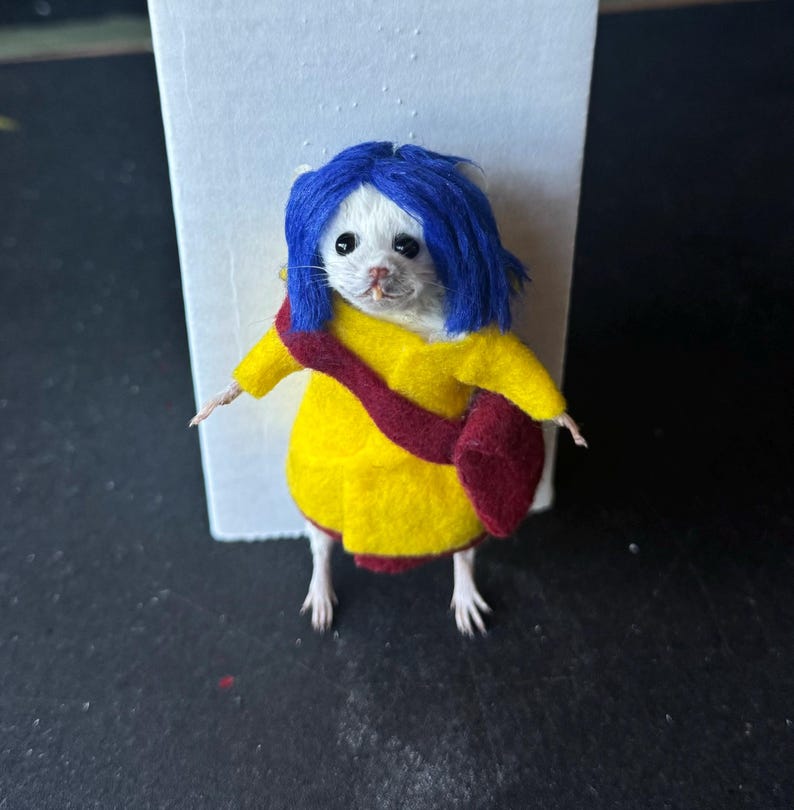 Puede incluir: Un rat&oacute;n blanco vestido con un abrigo amarillo con un bolso rojo y una peluca azul, que se asemeja a un personaje de una pel&iacute;cula. El rat&oacute;n est&aacute; de pie sobre sus patas traseras, con sus peque&ntilde;os brazos extendidos. El fondo es una tarjeta blanca y una superficie negra.