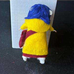 Puede incluir: Mu&ntilde;eca de fieltro hecha a mano con un abrigo amarillo con una faja y ribetes rojos. La mu&ntilde;eca tiene un sombrero azul y zapatos blancos. La mu&ntilde;eca est&aacute; de pie frente a un fondo blanco y una superficie negra.