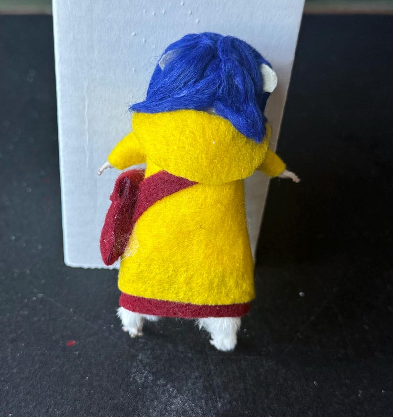 Puede incluir: Mu&ntilde;eca de fieltro hecha a mano con un abrigo amarillo con una faja y ribetes rojos. La mu&ntilde;eca tiene un sombrero azul y zapatos blancos. La mu&ntilde;eca est&aacute; de pie frente a un fondo blanco y una superficie negra.