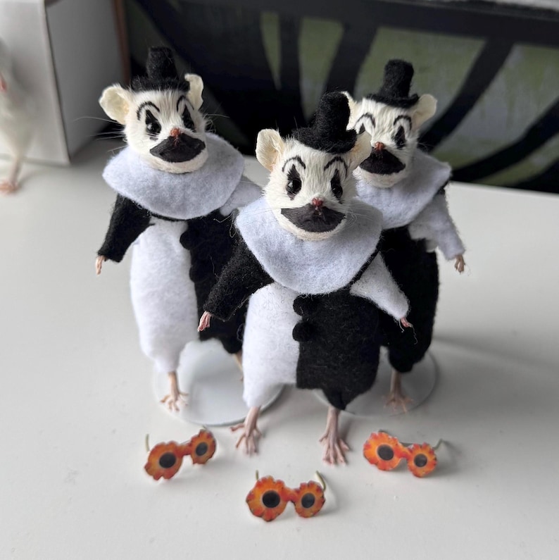May include: Three small mouse figurines dressed in black and white clown costumes, each wearing a black top hat and a white ruffled collar. The mice have painted facial features and are accompanied by three pairs of small orange flower-shaped glasses.