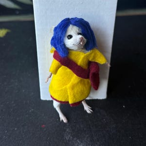 Puede incluir: Un rat&oacute;n blanco vestido con un abrigo de fieltro amarillo, una faja burdeos y una peluca azul. El rat&oacute;n est&aacute; posado sobre un fondo blanco y una superficie negra. El disfraz es una creaci&oacute;n caprichosa y artesanal.