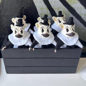 May include: A group of small white mouse figurines dressed as clowns, each wearing a black top hat, a white felt collar, and black clothing. The mice have painted black clown makeup around their eyes and mouths and are displayed in a black wooden crate.