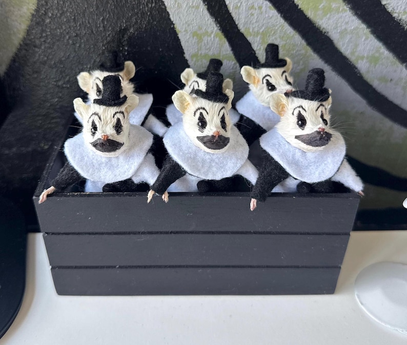 May include: A group of small white mouse figurines dressed as clowns, each wearing a black top hat, a white felt collar, and black clothing. The mice have painted black clown makeup around their eyes and mouths and are displayed in a black wooden crate.