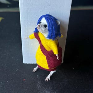 Puede incluir: Una figura de rat&oacute;n vestida con un abrigo amarillo, una bolsa roja y una peluca azul. El rat&oacute;n est&aacute; colocado frente a un fondo blanco. El rat&oacute;n lleva un abrigo amarillo con ribete rojo y una bolsa roja.