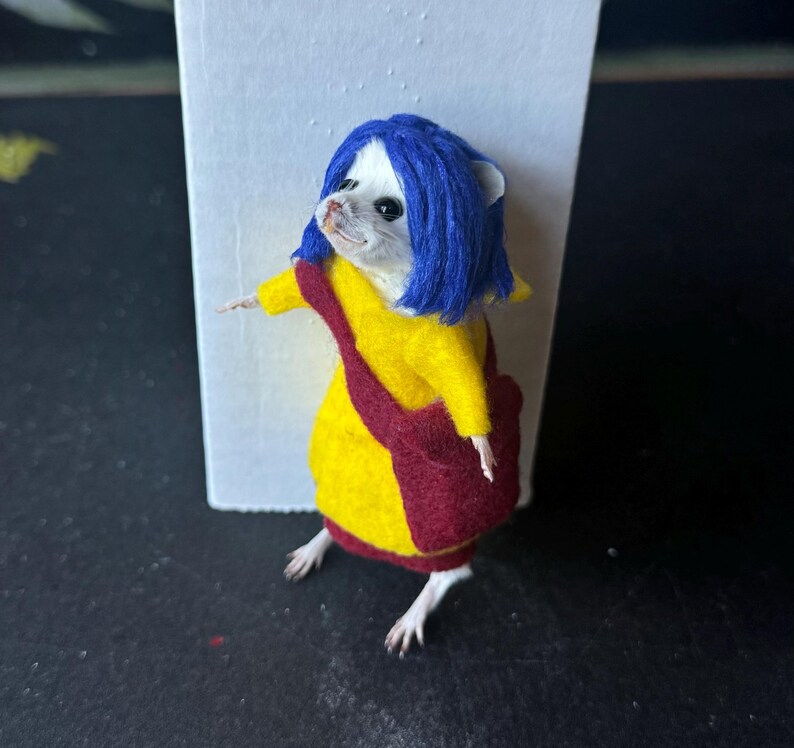 Puede incluir: Una figura de rat&oacute;n vestida con un abrigo amarillo, una bolsa roja y una peluca azul. El rat&oacute;n est&aacute; colocado frente a un fondo blanco. El rat&oacute;n lleva un abrigo amarillo con ribete rojo y una bolsa roja.