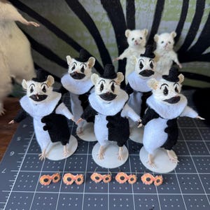May include: A group of six small mouse figurines dressed as clowns in black and white outfits with ruffled collars and tiny black hats. Each mouse has painted clown makeup on its face. Five small orange, circular accessories are placed in front.