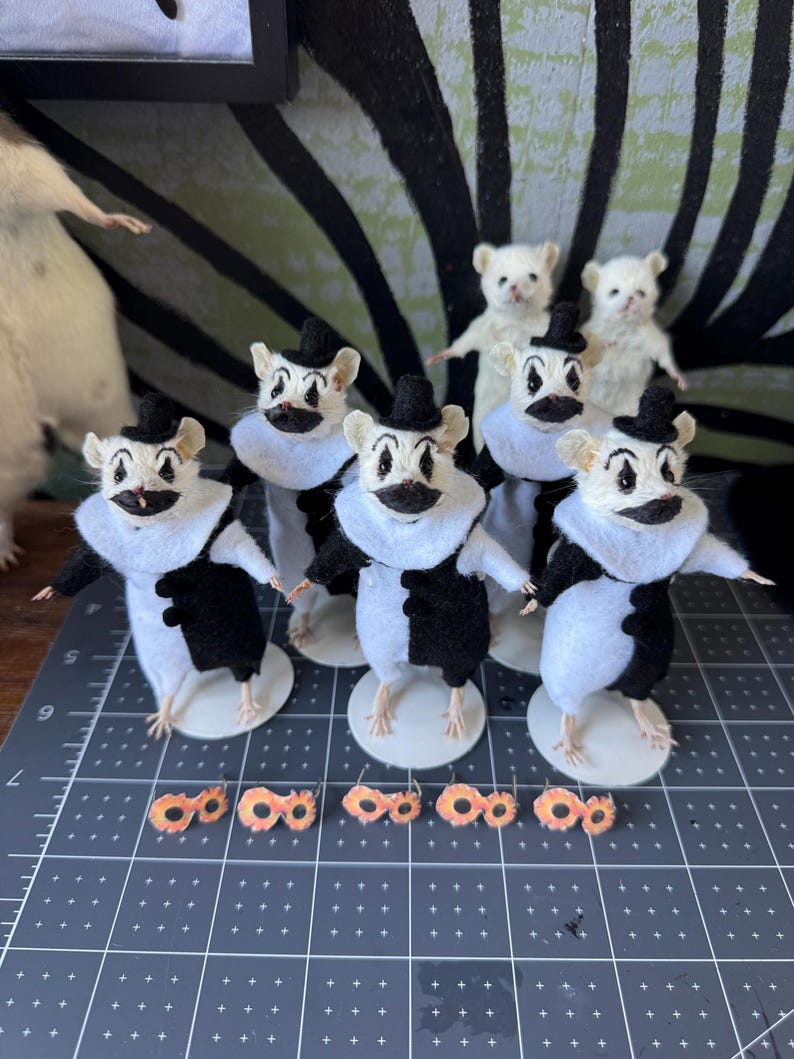May include: A group of six small mouse figurines dressed as clowns in black and white outfits with ruffled collars and tiny black hats. Each mouse has painted clown makeup on its face. Five small orange, circular accessories are placed in front.