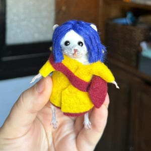 May include: A small needle-felted mouse figurine with bright blue hair, wearing a yellow coat with a dark red sash and matching red trim. The mouse has a white face, black bead eyes, and a small pink nose, standing upright with visible paws.