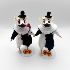 May include: Two taxidermy mice dressed as clowns, each wearing a black top hat and a white ruffled collar. One mouse has red blood-like splatters on its white and black outfit. Both figures stand on circular white bases with orange accents.