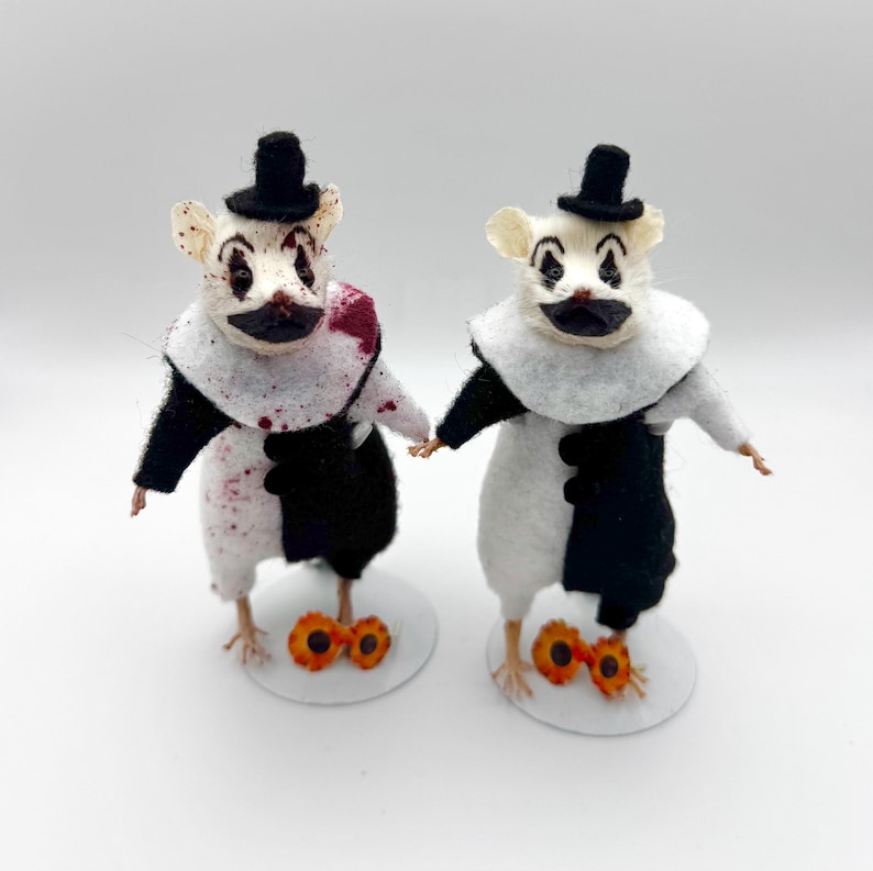 May include: Two taxidermy mice dressed as clowns, each wearing a black top hat and a white ruffled collar. One mouse has red blood-like splatters on its white and black outfit. Both figures stand on circular white bases with orange accents.