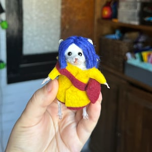Puede incluir: Un rat&oacute;n de fieltro hecho a mano vestido como un personaje de una pel&iacute;cula. El rat&oacute;n lleva un abrigo amarillo, un bolso rojo y una peluca azul. El rat&oacute;n est&aacute; sostenido en la mano de una persona.