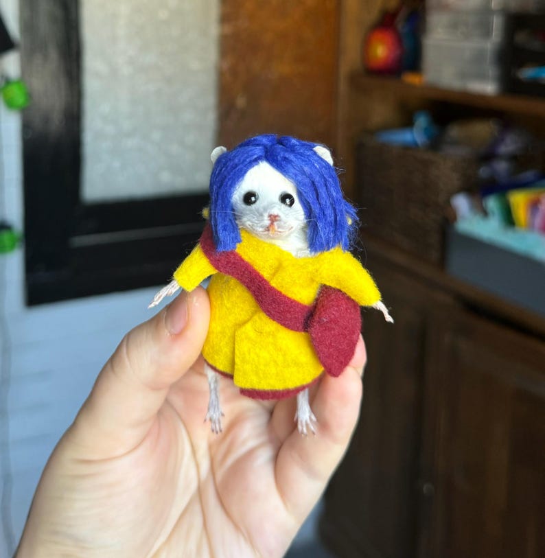 Puede incluir: Un rat&oacute;n de fieltro hecho a mano vestido como un personaje de una pel&iacute;cula. El rat&oacute;n lleva un abrigo amarillo, un bolso rojo y una peluca azul. El rat&oacute;n est&aacute; sostenido en la mano de una persona.
