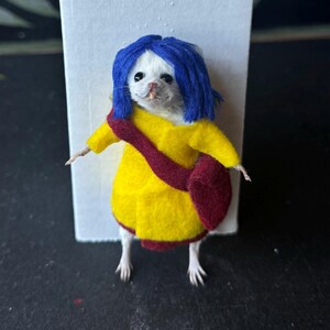 Puede incluir: Un rat&oacute;n blanco vestido con un abrigo amarillo, un bolso granate y una peluca azul. El rat&oacute;n est&aacute; de pie frente a un fondo blanco. El disfraz es una r&eacute;plica en miniatura de un personaje de una pel&iacute;cula.