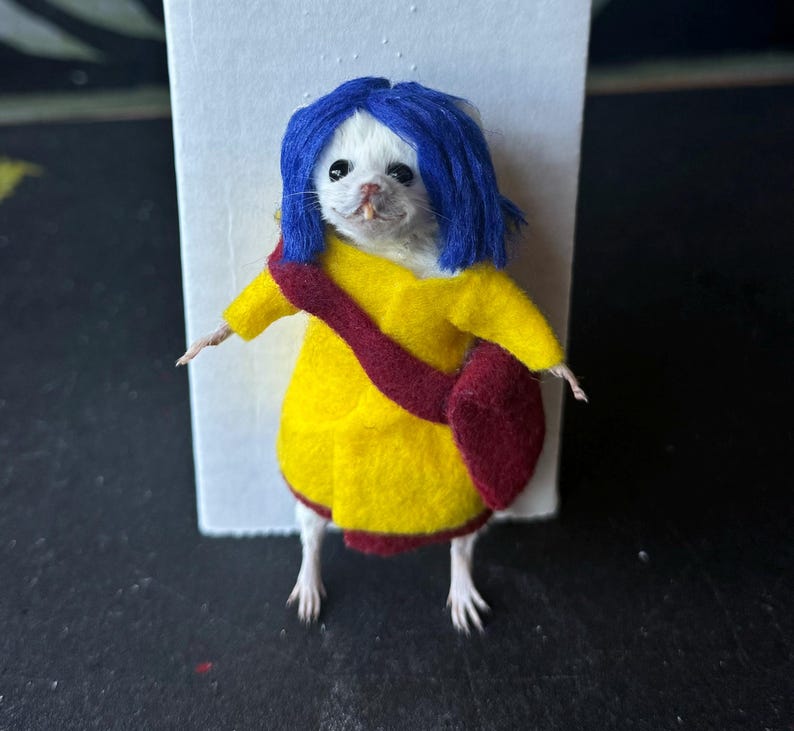 Puede incluir: Un rat&oacute;n blanco vestido con un abrigo amarillo, un bolso granate y una peluca azul. El rat&oacute;n est&aacute; de pie frente a un fondo blanco. El disfraz es una r&eacute;plica en miniatura de un personaje de una pel&iacute;cula.