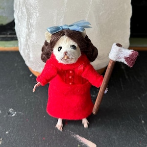 May include: A small, white mouse doll wearing a red dress and a blue bow. The doll is holding a small, white axe with a red stain on the blade.