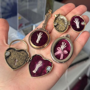 May include: A collection of four metal pendants featuring deep burgundy velvet inserts. Three pendants contain small white hand sculptures, one with a pink ribbon. The set includes a heart-shaped locket, a round mirror-style pendant, and two oval lockets.