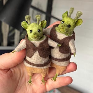 May include: Two small, handcrafted felt mouse figures with green, textured heads and upright, antenna-like ears. Each mouse wears a beige tunic with a dark brown waistcoat and a matching brown belt, finished with light tan lower bodies and thin feet.