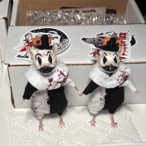 May include: Two small mouse figurines dressed as clowns, featuring white and black outfits, ruffled collars, and tiny black top hats. The mice have painted clown faces with red accents and splatters of red paint resembling blood on their clothing.