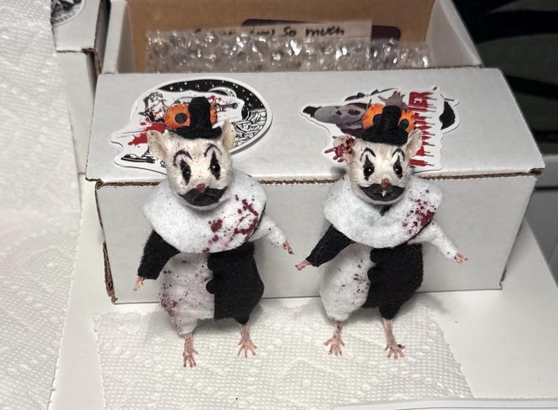 May include: Two small mouse figurines dressed as clowns, featuring white and black outfits, ruffled collars, and tiny black top hats. The mice have painted clown faces with red accents and splatters of red paint resembling blood on their clothing.
