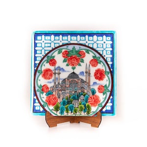 Può includere: Una piastrella di ceramica con un motivo geometrico blu e bianco e una piastrella rotonda e colorata che presenta una moschea con rose rosse intorno. Le piastrelle sono su un supporto in legno.