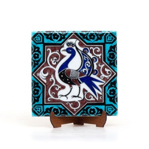 May include: A ceramic tile with a blue and white peacock design. The peacock is surrounded by intricate floral patterns in shades of brown and black. The tile is set against a turquoise background with a black border.