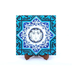 Op de afbeelding: Vierkante keramische tegel met een zonnegelaatontwerp in het midden, omgeven door een achtpuntig sterpatroon. De tegel heeft een kleurenpalet van wit, blauw en zwart. Het wordt tentoongesteld op een kleine houten standaard.