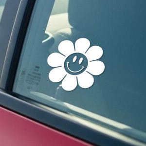 May include: A white daisy flower sticker with a smiling face is affixed to the rear window of a red car. The sticker is visible from the outside, with the car's interior and a portion of the dashboard visible through the glass.