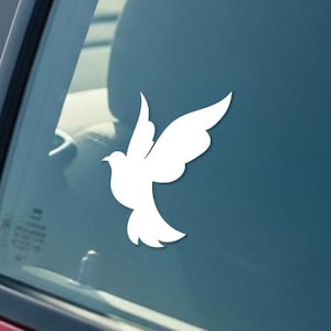 May include: A white dove decal is affixed to the window of a red car. The dove is depicted in a simple, stylized design, with its wings spread in flight. The car's interior is visible through the window.