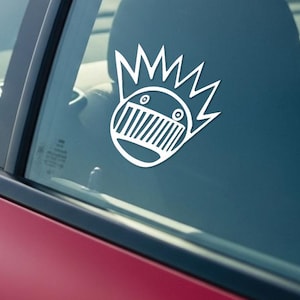 May include: White vinyl car decal featuring a stylized face with a crown-like design. The decal is applied to the rear window of a red car. The interior of the car is visible through the window.