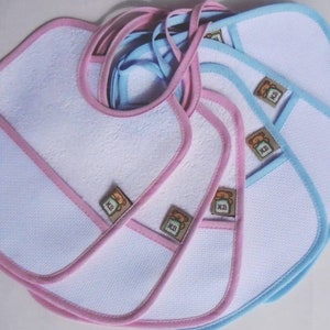 Puede incluir: Una pila de seis baberos blancos para bebé con ribetes rosas y azules. Cada babero tiene un bolsillo y un pequeño oso bordado con las letras "K.B."