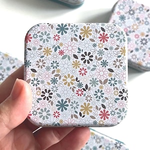 Può includere: Scatola quadrata con motivo floreale. Il design presenta piccoli fiori nei toni del rosso, marrone, giallo e blu su sfondo bianco. La scatola è tenuta in mano, con altre scatole simili visibili sullo sfondo.