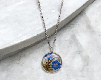 May include: A silver chain necklace with a round pendant featuring pressed blue and white flowers and fern leaves encased in clear resin.