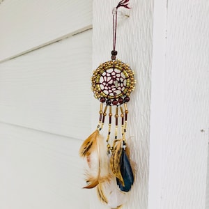 May include: A small dream catcher with brown and gold beads, a burgundy web, and brown, white, and black feathers.