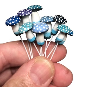 May include: A collection of miniature blue and white polka dot mushrooms with white stems. The mushrooms are arranged in a hand.