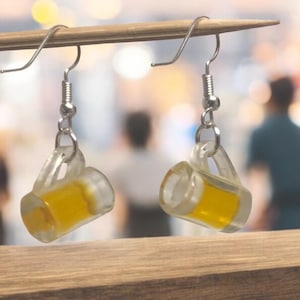 Può includere: Un paio di orecchini d'argento con ciondoli a forma di boccale di birra in vetro trasparente. I boccali sono riempiti di un liquido giallo che ricorda la birra.