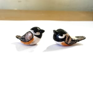 以下が含まれることがあります： 2つの小さなセラミック製の鳥の置物。 各鳥は黒い頭、白い頬、茶色とオレンジ色の体を持っています。 鳥は並んでおり、白い表面に置かれています。 置物は詳細で、手作りのように見えます。