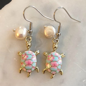 Może przedstawiać: Para złotych kolczyków w kształcie żółwia z jasnoniebieskim i różowym wzorem kwiatowym. Każdy kolczyk ma zawieszkę z białą perłą.