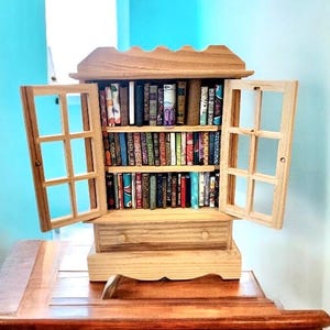 Puede incluir: Una estantería de madera en miniatura con puertas abiertas, llena de pequeños libros de varios colores y tamaños. La estantería tiene un cajón y una parte superior decorativa. Las puertas tienen un diseño de vidriera. La estantería está sobre una mesa de madera.