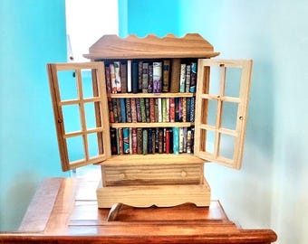 May include: A miniature wooden bookcase with open doors, filled with tiny books of various colors and sizes. The bookcase has a drawer and a decorative top. The doors have a window-pane design. The bookcase is sitting on a wooden table.