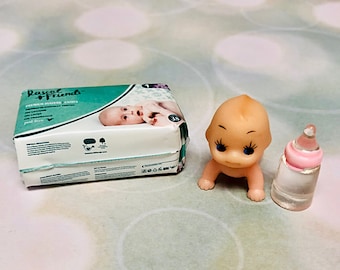May include: A package of Rascal + Friends premium diapers in size 1, a small peach-colored plastic baby doll with blue eyes and long eyelashes, and a miniature clear plastic baby bottle with a pink cap and a clear nipple top.
