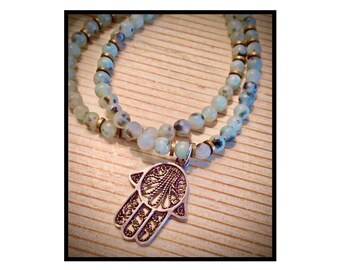 Puede incluir: Un collar de cuentas con un colgante de mano de Hamsa plateado. El collar presenta cuentas de color azul claro y blanco con espaciadores dorados. El colgante de mano de Hamsa tiene un diseño de filigrana intrincado.