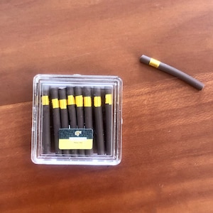 Puede incluir: Una caja de plástico transparente que contiene varios objetos cilíndricos de color marrón oscuro con bandas doradas. Un objeto similar descansa fuera de la caja. Los objetos se asemejan a cigarros. Una pequeña etiqueta con texto es visible en la caja.