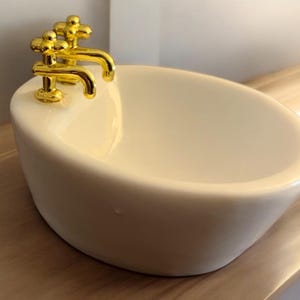 Puede incluir: Un lavabo de cerámica blanca con un grifo dorado. El lavabo tiene forma ovalada y un acabado liso y brillante.